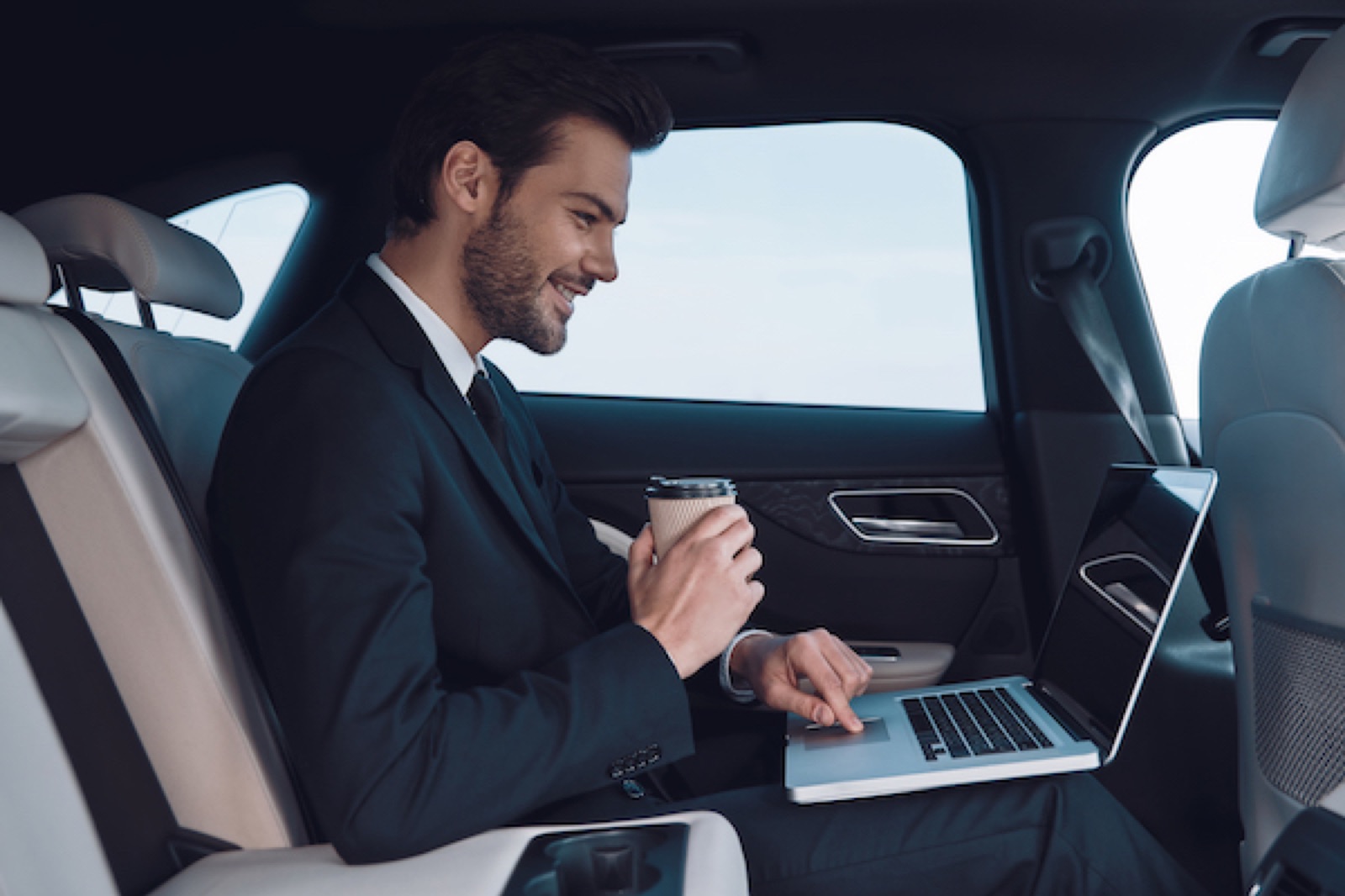 Passenger working on a laptop with coffee in the back of a chauffeur car